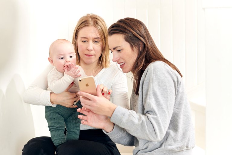 Ung kvinne holder en baby mens hun og venninne ser på en smarttelefon. Illustrasjonsbilde